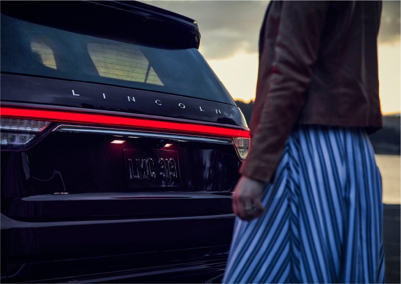 A person is shown near the rear of a 2023 Lincoln Aviator® SUV as the Lincoln Embrace illuminates the rear lights | Gary Yeomans Lincoln Ocala in Ocala FL